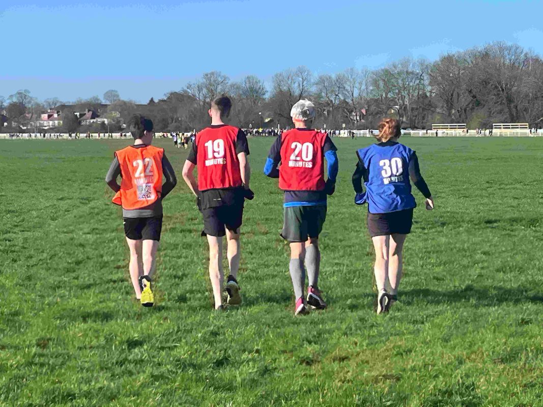 Four parkrun pacers wearing numbered bibs (22, 19, 20, and 30 minutes) running together across an open grass field at York parkrun, with trees and houses visible in the background under a clear blue sky.