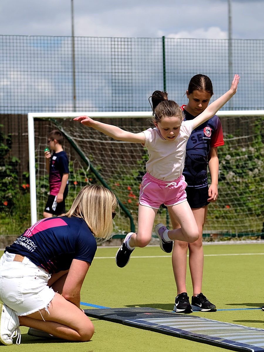 A girl long jumping into the sandpit with a Vale of York coach alongside